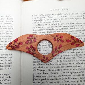Puede incluir: Un marcap&aacute;ginas de madera con una lupa y un dise&ntilde;o floral rojo y marr&oacute;n. El marcap&aacute;ginas est&aacute; sobre una p&aacute;gina de un libro con el texto "Jane Eyre" visible.