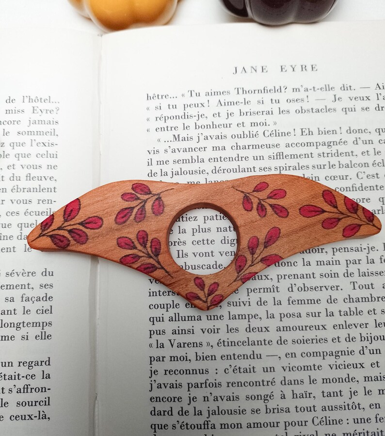 Puede incluir: Un marcap&aacute;ginas de madera con una lupa y un dise&ntilde;o floral rojo y marr&oacute;n. El marcap&aacute;ginas est&aacute; sobre una p&aacute;gina de un libro con el texto "Jane Eyre" visible.