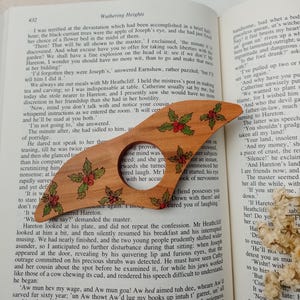May include: A wooden bookmark with a holly design. The bookmark is shaped like a Christmas tree and has red berries and green leaves. The bookmark is resting on a page of a book with the text "Wuthering Heights" visible.