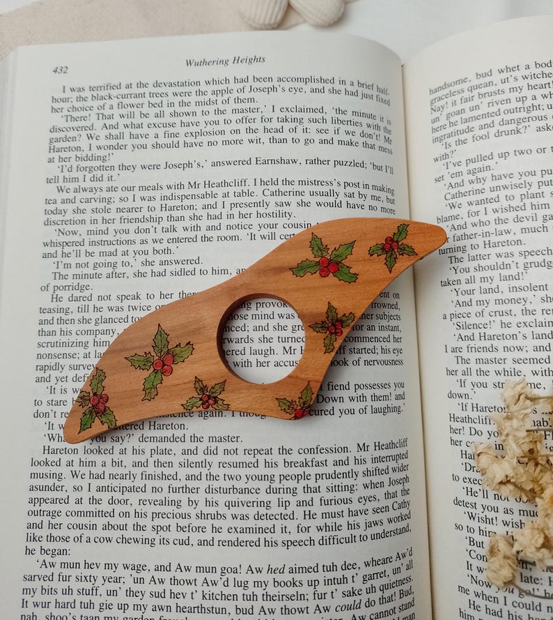 May include: A wooden bookmark with a holly design. The bookmark is shaped like a Christmas tree and has red berries and green leaves. The bookmark is resting on a page of a book with the text "Wuthering Heights" visible.