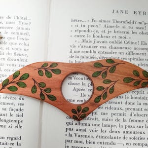 Puede incluir: Un marcap&aacute;ginas de madera con una lupa. El marcap&aacute;ginas est&aacute; decorado con hojas verdes y tiene una veta de madera marr&oacute;n. La lupa muestra una parte de una p&aacute;gina de libro con el texto "m'&eacute;coutiez chose la pl Apr&egrave;s ce Ils v - une emb"