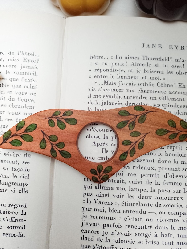 Puede incluir: Un marcap&aacute;ginas de madera con una lupa. El marcap&aacute;ginas est&aacute; decorado con hojas verdes y tiene una veta de madera marr&oacute;n. La lupa muestra una parte de una p&aacute;gina de libro con el texto "m'&eacute;coutiez chose la pl Apr&egrave;s ce Ils v - une emb"