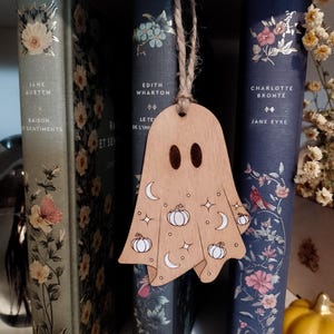 Pode incluir: Um marcador de livros em forma de fantasma de madeira com uma corda, decorado com abóboras, luas e estrelas. O marcador de livros está pendurado em frente a uma fila de livros antigos com desenhos florais e títulos como "Jane Austen" e "Jane Eyre."
