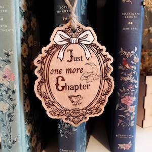 Peut inclure: Marque-page en bois avec l'inscription "Just one more Chapter" dans un cadre décoratif, suspendu devant une rangée de livres. Le marque-page présente un nœud, une lune, des nuages et une tasse à thé. Les livres ont des motifs floraux.