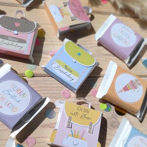 Könnte beinhalten: Eine Sammlung kleiner rechteckiger Riegel, verpackt in dekorativem Papier mit schulbezogenen Illustrationen wie Schulranzen, Federmäppchen und Texten wie Meine Einschulung, Hurra! Endlich Schulkind und Ich bin erste Klasse.