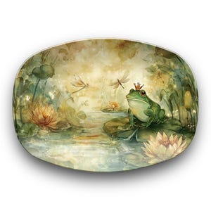 Può includere: Piatto decorativo ovale con un'illustrazione ad acquerello di una rana con una corona, seduta su una ninfea. La scena comprende libellule, ninfee e uno stagno, con una tavolozza di colori morbidi e sognanti.