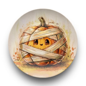 May include: A round decorative plate featuring a cartoon pumpkin wrapped in bandages, with a smiling face. The pumpkin is orange with brown bandages, set against a light beige background with autumn leaves. A fun Halloween design.
