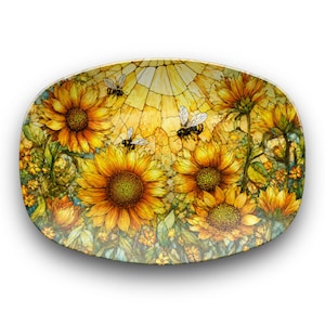 Può includere: Un piatto decorativo di forma ovale caratterizzato da una vivace illustrazione di girasoli gialli e api su uno sfondo in stile vetrata. Il design incorpora tonalità oro, arancione e verde, con petali e foglie dettagliati.