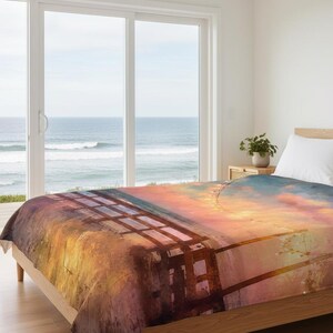 May include: A bed with a blanket featuring a watercolor painting of a sunset over the ocean. The blanket has a warm color palette of orange, yellow, and purple, with a wooden fence in the foreground.