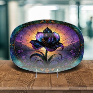 May include: An oval decorative plate with a stylized flower design. The flower is in shades of purple and blue against a sunset backdrop. The plate has a teal and gold border and is displayed on a clear stand.