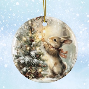 Puede incluir: Adorno navideño redondo de cerámica con una ilustración de acuarela de un conejito con bufanda, alcanzando una luz brillante en un árbol de Navidad nevado. El adorno tiene una cuerda dorada para colgar.