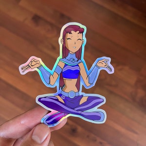 May include: A holographic sticker featuring a cartoon character with red hair, a blue and purple outfit, and a serene expression. The character is sitting in a meditative pose with their hands in a mudra.