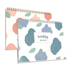 May include: Two spiral-bound weekly planners. The covers feature a white background with abstract shapes in coral, blue, and green. The front planner has the words "Weekly planner" in a white rectangle.