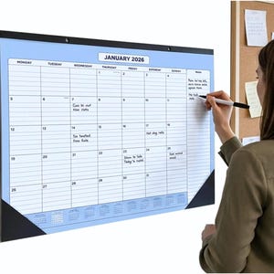 May include: A large desk calendar for January 2026, with a blue border and white grid. Someone is writing on the calendar with a black marker. The calendar is mounted on a wall. The days of the week are across the top.