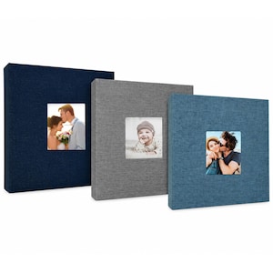 May include: Three square photo albums with fabric covers in navy blue, light gray, and denim blue. Each album features a centered square window displaying a different photograph: a couple embracing, a baby, and a couple posing together outdoors.