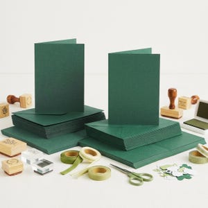 May include: Two stacks of dark green folded cards and matching envelopes, accompanied by several wooden rubber stamps, two rolls of green ribbon, a pair of metal scissors, a small ink pad, and a collection of small green paper cutouts.