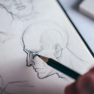 May include: A close-up of a hand drawing a portrait of a person's face in pencil on a white sheet of paper. The drawing is in progress, with the pencil being used to shade the nose and cheek area.