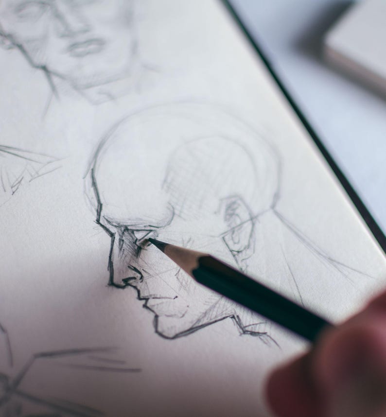 May include: A close-up of a hand drawing a portrait of a person's face in pencil on a white sheet of paper. The drawing is in progress, with the pencil being used to shade the nose and cheek area.
