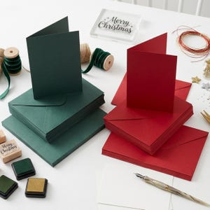 May include: A set of dark green and red folded cards with matching envelopes, accompanied by a clear acrylic stamp with the text Merry Christmas, green ribbon spools, ink pads, and a fountain pen on a white surface.