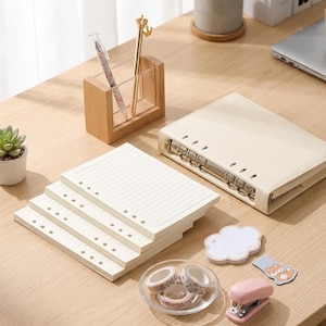 May include: A desk arrangement with stationery and office supplies. Includes a cream binder, lined paper, decorative tape, a stapler, and a wooden pen holder with pens. A small succulent plant and a cloud-shaped sticky note are also visible.