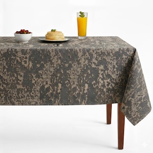 May include: A rectangular tablecloth with a gray and beige abstract pattern covers a wooden table. A white bowl of red berries, a stack of pancakes, and a glass of orange juice with a mint sprig are on the table.