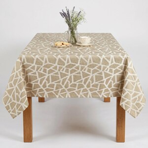 May include: A beige tablecloth with a white geometric pattern covers a wooden table. A glass jar with flowers and a teacup with a saucer sit on the table. A small plate of cookies is also on the table.