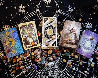 May include: Five tarot cards laid out on a black fabric background with a silver and gold star pattern. The cards are surrounded by small, colorful stones. The cards are: The Star, The Emperor, The World, The Sun, and The Moon.