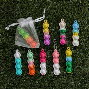 May include: A set of nine colourful pop it fidget toys, each with four bubbles. The toys are arranged in a row on a green grassy surface. The toys are made of silicone and have a variety of colours, including blue, green, yellow, orange, pink, and purple.