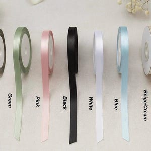 May include: Spools of satin ribbon in various colors, including brown, green, pink, black, white, blue, and beige/cream. Each spool is labeled with its color name. The ribbons are displayed against a neutral background.