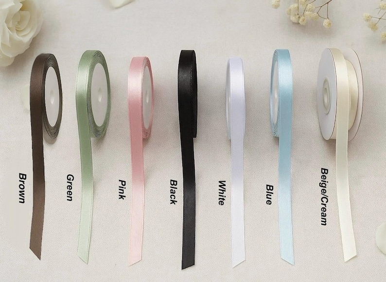 May include: Spools of satin ribbon in various colors, including brown, green, pink, black, white, blue, and beige/cream. Each spool is labeled with its color name. The ribbons are displayed against a neutral background.