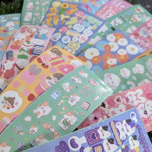 May include: A collection of colorful sticker sheets featuring various designs. The stickers showcase illustrations of animals, food, and floral patterns in shades of pink, blue, green, and yellow. The sheets are arranged on a green surface.