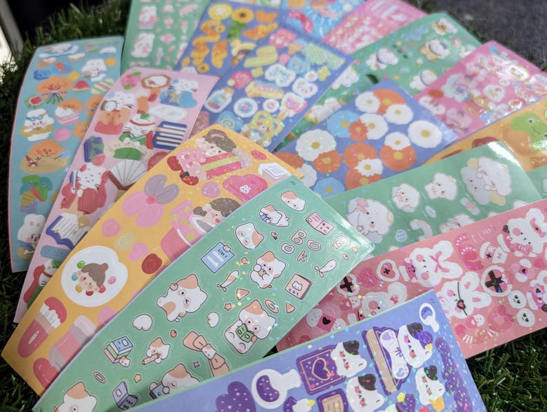 May include: A collection of colorful sticker sheets featuring various designs. The stickers showcase illustrations of animals, food, and floral patterns in shades of pink, blue, green, and yellow. The sheets are arranged on a green surface.