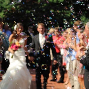 May include: A wedding ceremony with a bride in a white gown and a groom in a black suit walking down an aisle. Bubbles float in the air, and guests line the sides. The scene is outdoors with a blurred green background.