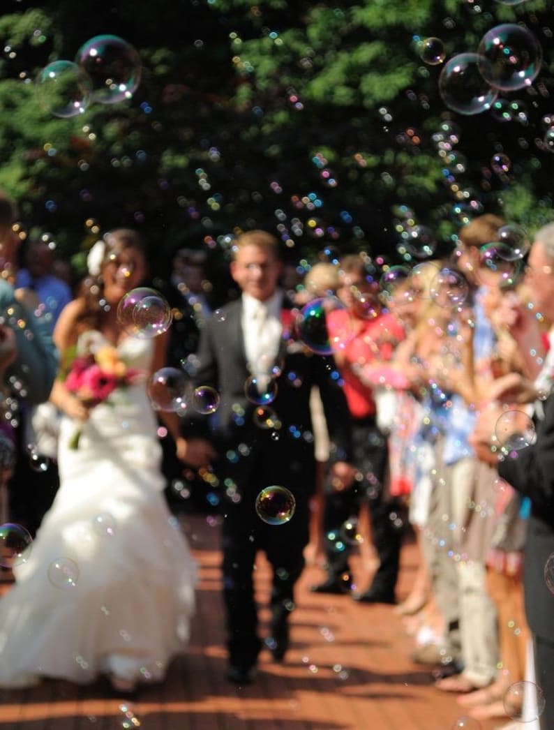 May include: A wedding ceremony with a bride in a white gown and a groom in a black suit walking down an aisle. Bubbles float in the air, and guests line the sides. The scene is outdoors with a blurred green background.