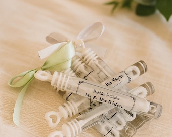 May include: A collection of clear bubble wands with heart-shaped handles and labels that read "Bubbles & Wishes Mr & Mrs Waleer" and "Mar Maynes". The wands are tied with white and green ribbons, arranged on a cream-colored surface.
