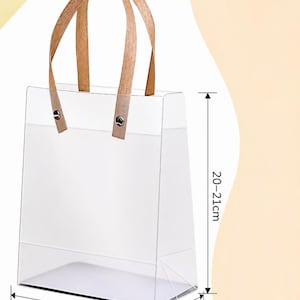 May include: Clear rectangular tote bag with wooden handles. The bag is 15cm (5.9in) wide, 7-8cm deep, and 20-21cm (7.9-8.3in) tall. The handles are attached with metal rivets. The bag is empty and sits on a light background.
