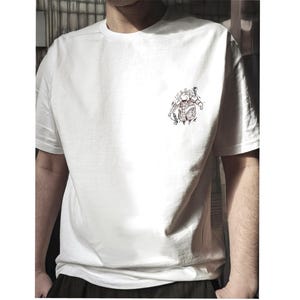 May include: A white short-sleeved t-shirt featuring a small, black-ink line art illustration on the upper left chest. The graphic depicts a stylised, round-bodied creature with spiky hair, wearing a patterned vest and holding a small object.