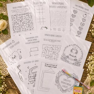 May include: A collection of wedding activity sheets, including an I Spy game, word search, and maze, are arranged on a wooden surface. A box of crayons and a multi-colored pen are also present. The sheets feature various wedding-themed illustrations and text.