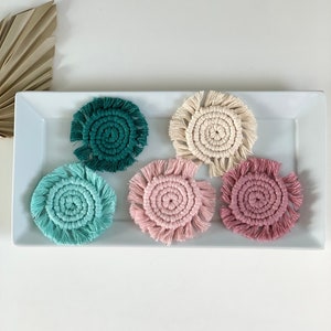 May include: Five handmade crocheted coasters in a variety of colors, including teal, pink, cream, and green. The coasters are displayed on a white rectangular plate.
