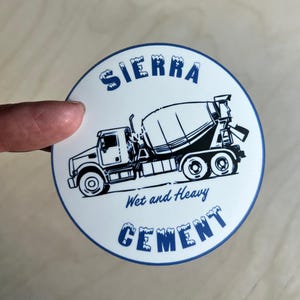 May include: Round white coaster with a blue border featuring a black and white illustration of a cement truck. The text "SIERRA Wet and Heavy CEMENT" is printed in blue, with a stylized font.