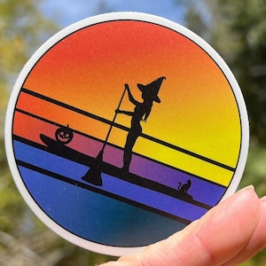 May include: A round sticker with a retro sunset design featuring a silhouette of a witch paddleboarding with a pumpkin and a cat on the board.