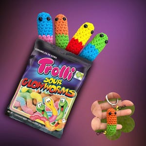 Puede incluir: Una bolsa de caramelos Trolli Sour Glowworms con cuatro juguetes de gusanos de ganchillo coloridos. Los gusanos son azules, naranjas, verdes y amarillos. También se muestra un llavero de ganchillo con forma de gusano. El fondo es un degradado púrpura.