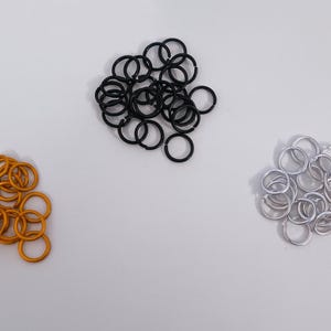 May include: Three piles of metal jump rings in gold, black, and silver. The jump rings are small, circular, and appear to be made of metal. The gold rings are clustered on the left, black in the middle, and silver on the right.