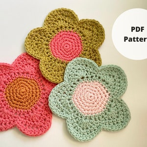 Peut inclure: Trois dessous-de-verre en forme de fleur crochetés dans différentes couleurs. Les dessous-de-verre sont réalisés avec un motif de granny square et ont un cercle central de couleur contrastante. Les couleurs sont jaune moutarde, rose corail et vert clair. Le texte "PDF Pattern" est visible en arrière-plan.
