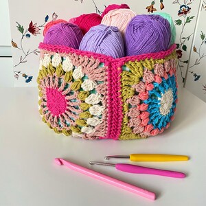 May include: A crocheted basket with a colorful granny square pattern holds balls of yarn in shades of pink, purple, and green. Two crochet hooks are also pictured.