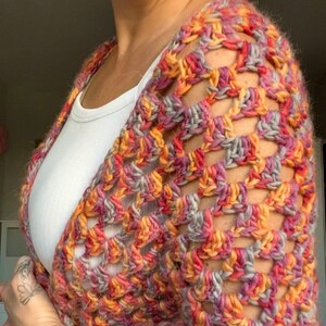 Pu&ograve; includere: Uno scialle all'uncinetto multicolore nei toni dell'arancione, del rosa e del grigio. Il capo fatto a mano &egrave; drappeggiato su una canotta bianca, mettendo in mostra il filato strutturato e il design aperto. I colori vivaci dello scialle creano un'estetica calda e invitante.