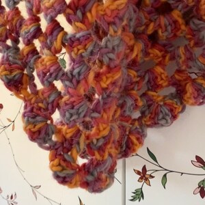 Pu&ograve; includere: Primo piano di uno scialle all'uncinetto fatto a mano. Lo scialle presenta un complesso design traforato nei toni dell'arancione, del viola e del grigio. Il filato appare spesso e strutturato, suggerendo un accessorio caldo e confortevole. L'estetica generale &egrave; rustica e artigianale.