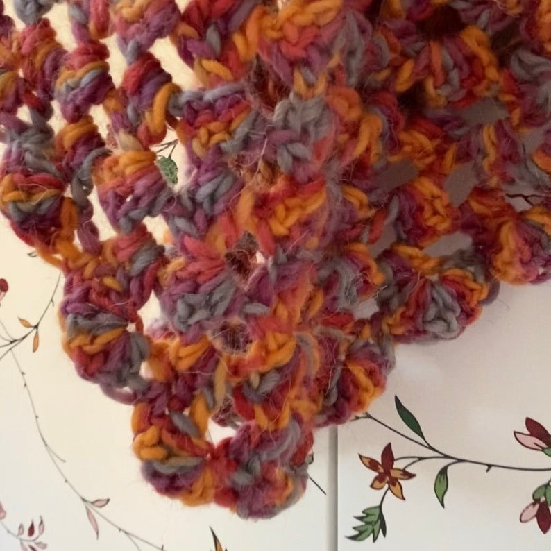 Pu&ograve; includere: Primo piano di uno scialle all'uncinetto fatto a mano. Lo scialle presenta un complesso design traforato nei toni dell'arancione, del viola e del grigio. Il filato appare spesso e strutturato, suggerendo un accessorio caldo e confortevole. L'estetica generale &egrave; rustica e artigianale.