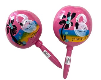 May include: A pair of pink maracas with painted designs of flamingos and palm trees. The maracas have the word "Mexico" written on them.