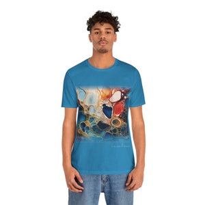 May include: A turquoise blue t-shirt with a colorful abstract design featuring swirling shapes and colors. The design is reminiscent of liquid paint or a galaxy. The text "Colour Rivers" is printed on the bottom right corner of the design.